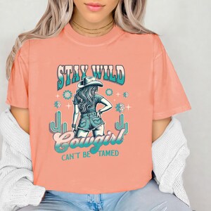 May include: A peach-coloured t-shirt with a graphic of a woman wearing a cowboy hat and jeans. The text "Stay Wild Cowgirl Can't Be Tamed" is printed on the shirt.