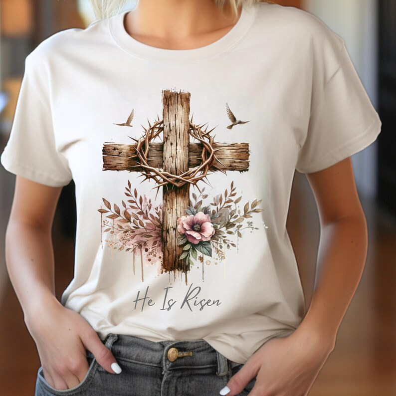 May include: Cream-colored t-shirt featuring a detailed graphic of a wooden cross adorned with a crown of thorns and floral elements. Two birds are depicted on either side of the cross. The phrase "He Is Risen" is written in script below the cross.