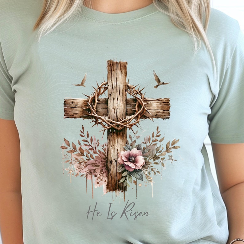May include: A light green t-shirt featuring a watercolor design of a wooden cross with a crown of thorns, flowers, and the text "He Is Risen". Two birds are flying above the cross. The shirt is a soft, comfortable material.