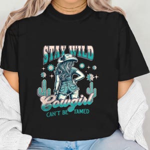 May include: Black t-shirt featuring a graphic of a cowgirl wearing a hat and jeans. The text "Stay Wild Cowgirl Can't Be Tamed" is printed on the shirt.