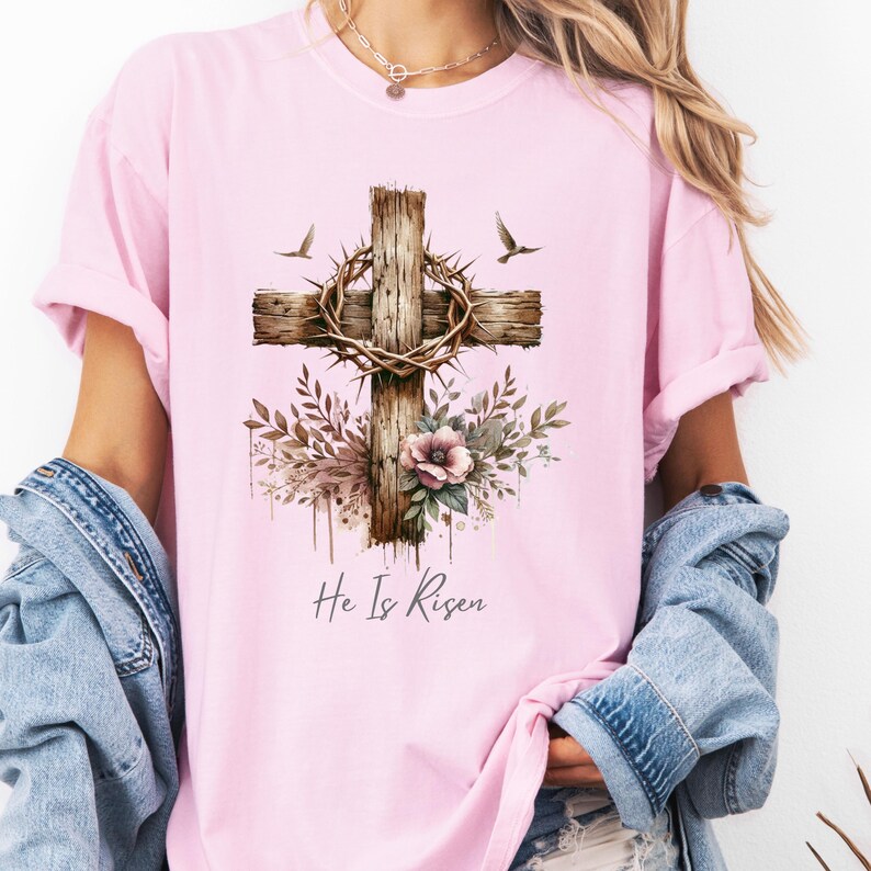 May include: Pink t-shirt featuring a watercolor design of a wooden cross with a crown of thorns, floral accents, and two birds. The text "He Is Risen" is written below the cross.