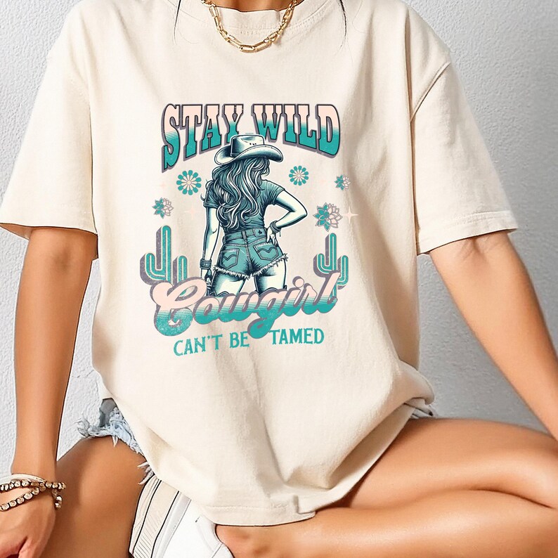 May include: A beige t-shirt featuring a graphic print of a woman wearing a cowboy hat and jeans. The text "Stay Wild Cowgirl Can't Be Tamed" is printed on the shirt.