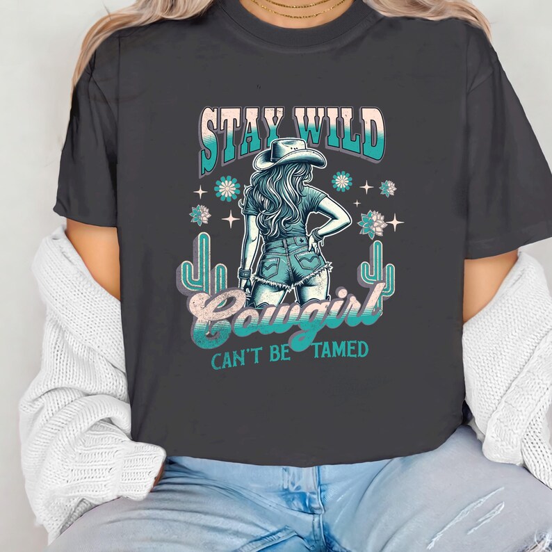 May include: A dark grey t-shirt with a graphic print of a woman wearing a cowboy hat and jeans. The text "Stay Wild Cowgirl Can't Be Tamed" is printed on the shirt.