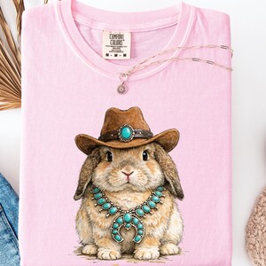 May include: A light pink Comfort Colors t-shirt with a graphic of a rabbit wearing a brown cowboy hat and turquoise jewelry. A delicate necklace with a flower pendant is draped across the shirt.