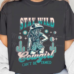 May include: A dark grey t-shirt with a graphic design featuring a woman wearing a cowboy hat and jeans. The text "Stay Wild Cowgirl Can't Be Tamed" is printed on the shirt.
