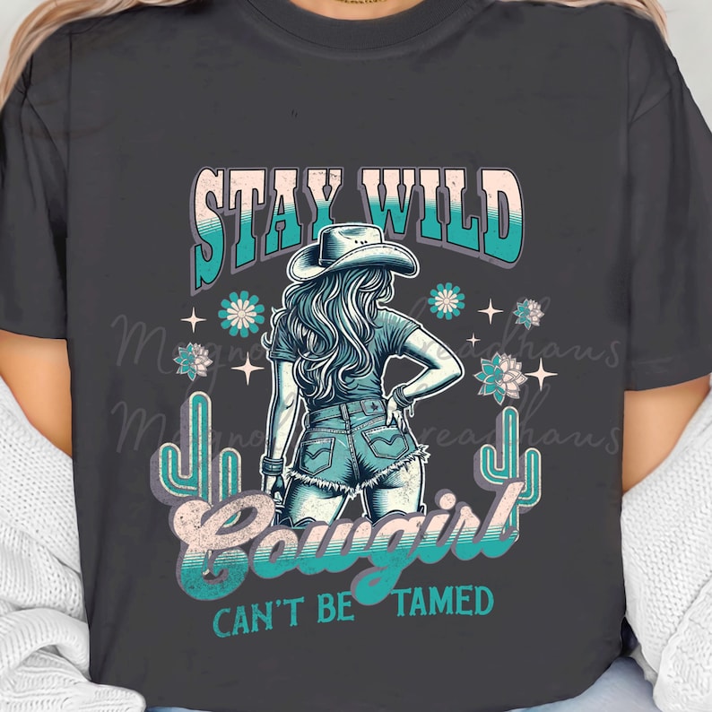 May include: A dark grey t-shirt with a graphic design featuring a woman wearing a cowboy hat and jeans. The text "Stay Wild Cowgirl Can't Be Tamed" is printed on the shirt.