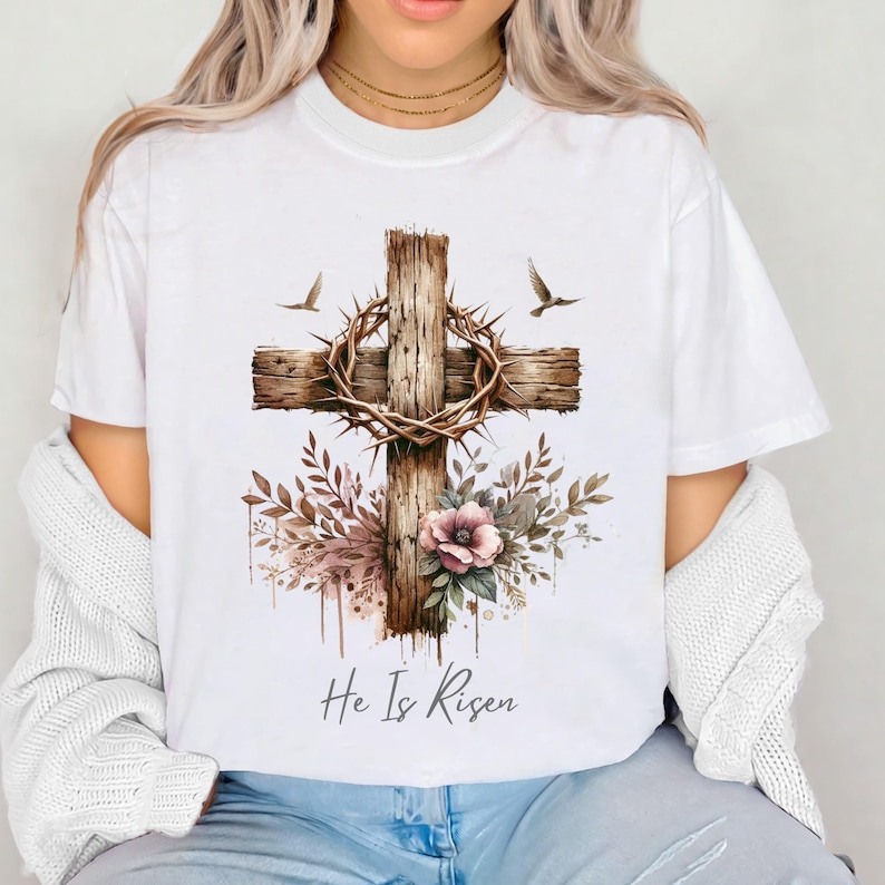 May include: White t-shirt featuring a watercolor design of a wooden cross with a crown of thorns, flowers, and two birds. The text "He Is Risen" is printed below the cross. Religious-themed apparel.