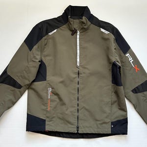 Może przedstawiać: Zielono-oliwkowa i czarna kurtka RLX Expedition z pełnym zamkiem błyskawicznym i stójką. Kurtka posiada czarne panele na ramionach i rękawach, z białymi i czerwonymi akcentami. Logo RLX jest widoczne na rękawie.