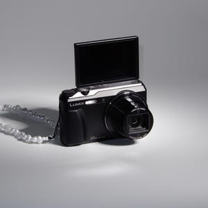 May include: A black digital camera with a silver lens and a flip-up screen. The camera has a black strap with white beads.