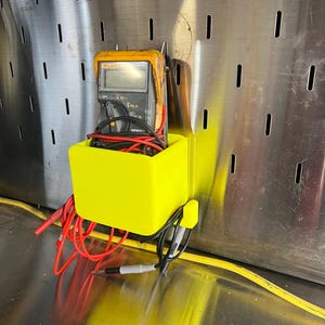 May include: A yellow plastic wall-mounted holder with a digital multimeter and red and black wires inside. The holder is attached to a metal pegboard.