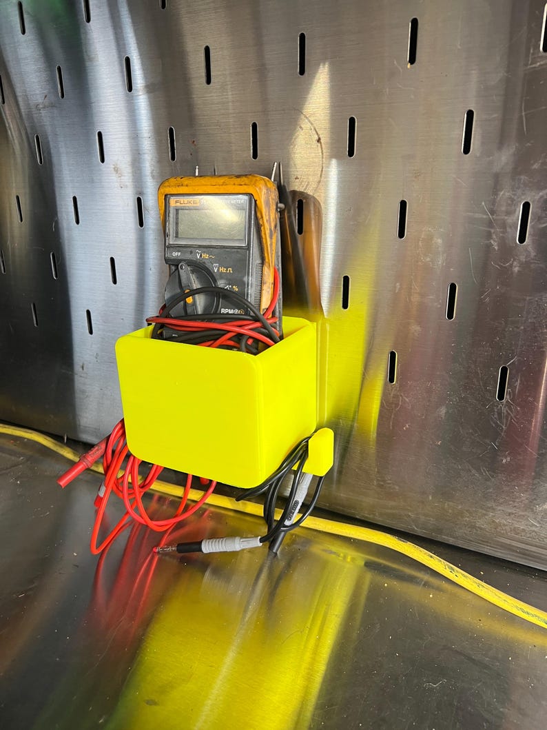 May include: A yellow plastic wall-mounted holder with a digital multimeter and red and black wires inside. The holder is attached to a metal pegboard.