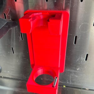 May include: A bright red plastic wall mount with a circular opening and a small metal rod extending from the bottom. The mount is designed to hold a tool or other object.