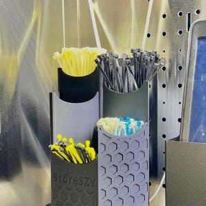 May include: A wall-mounted organizer with four compartments, each holding a different color of cable ties. The organizer is made of black and gray plastic. The bottom left compartment has the text "StoreSZY".