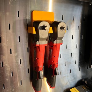 May include: Two red and black impact wrenches are hanging on a wall mounted tool organizer. The organizer is made of orange plastic and has two slots for the wrenches. The wrenches are hanging vertically with the handles pointing down.