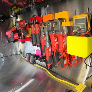 May include: A collection of red and black power tools are organized on a metal wall with orange and yellow plastic holders. The tools include impact drivers, wrenches, and a multimeter.