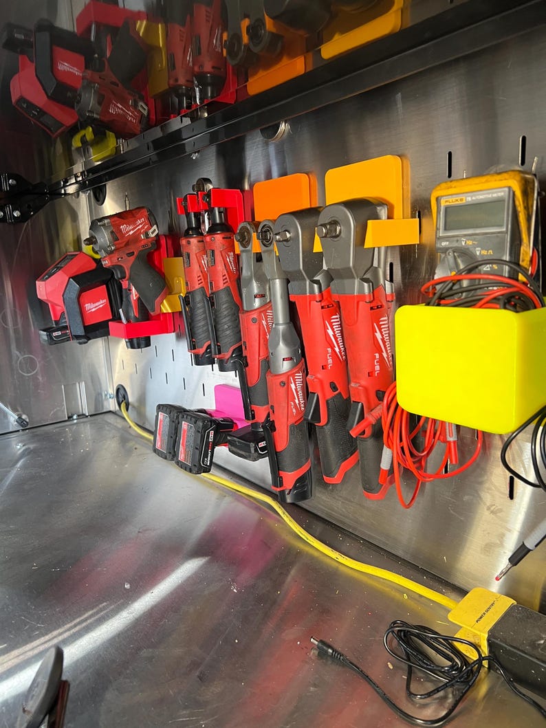 May include: A collection of red and black power tools are organized on a metal wall with orange and yellow plastic holders. The tools include impact drivers, wrenches, and a multimeter.