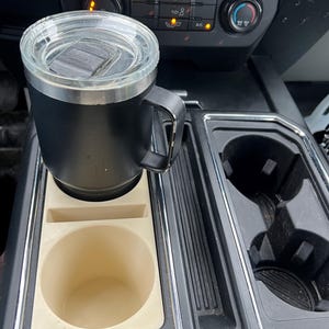 以下が含まれることがあります： 車のセンターコンソール用の白いプラスチック製のカップホルダーインサート。インサートにはカップホルダーが2つと小さな収納スペースがあります。蓋付きの黒いトラベルマグがカップホルダーの1つに入っています。