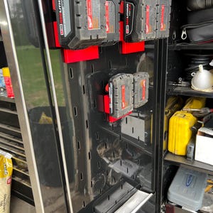 May include: A black metal tool storage cabinet with a pegboard wall. The pegboard has several red and black battery packs hanging on it. The batteries are labeled "18V LITHIUM" and "20V MAX".