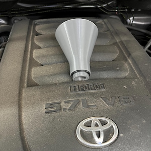 May include: A gray plastic funnel sits on top of a black engine compartment. The engine compartment has a silver Toyota emblem and the text "5.7L V8" and "TRD FORCE".