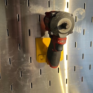 May include: A yellow wall mount with a red and black impact driver attached to it. The wall mount is attached to a metal pegboard.