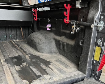 May include: The rear of a black pickup truck's load bed with a fishing rod and reel hanging from a red rod holder. The truck bed is lined with a black carpet and has a small pink air freshener hanging from the rearview mirror.