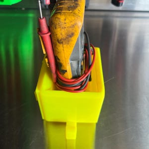 May include: A yellow and black multimeter with red and black test leads is stored in a bright yellow plastic holder.