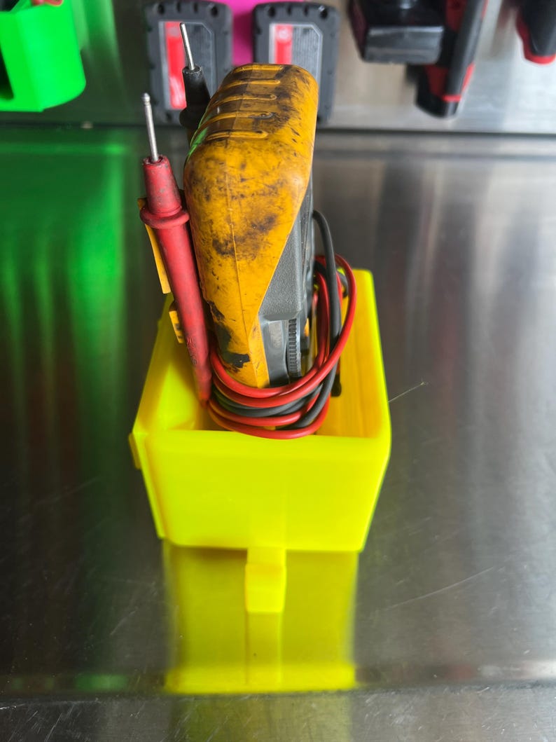 May include: A yellow and black multimeter with red and black test leads is stored in a bright yellow plastic holder.