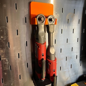 May include: Two red and black cordless impact wrenches are hanging on an orange wall mount. The mount is attached to a silver pegboard.