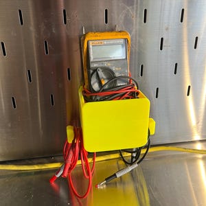 May include: A yellow plastic holder with a black Fluke 75 automotive meter inside. The meter has red and black test leads attached to it. The holder is mounted on a pegboard.