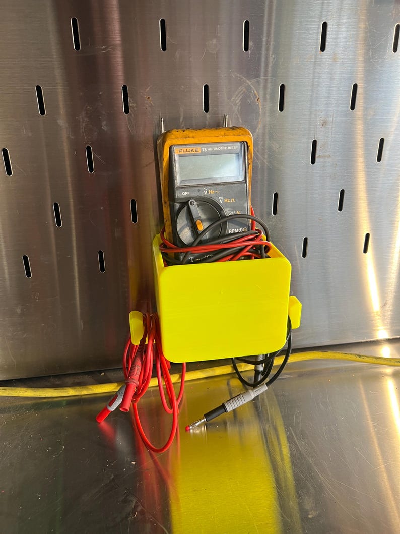 May include: A yellow plastic holder with a black Fluke 75 automotive meter inside. The meter has red and black test leads attached to it. The holder is mounted on a pegboard.