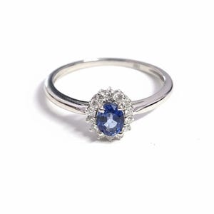 May include: A silver ring featuring a central oval-cut blue gemstone, surrounded by a halo of smaller, clear stones. The ring's band is thin and polished, creating a classic and elegant design. The ring is set against a white background.