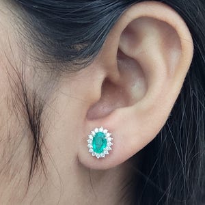 Op de afbeelding: Ovale smaragd oorknopjes met een halo van kleine, ronde diamanten. De smaragd heeft een levendige groene kleur en de diamanten schitteren. De oorbellen zijn gezet in een wit metaal.