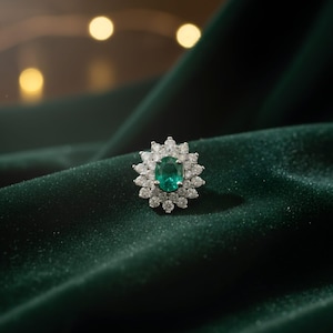 May include: A close-up of a gemstone ring featuring an oval-cut emerald center stone surrounded by a halo of small, round diamonds. The ring is set on a dark green velvet surface, creating a luxurious and elegant aesthetic.