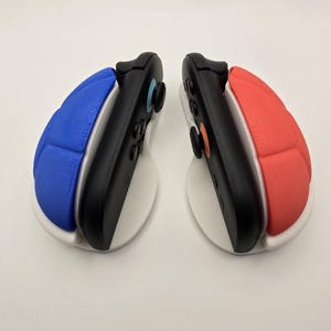 May include: Two black game controllers with blue and red padded grips, resting on white stands. The controllers have buttons in various colors. The setup is on a white surface.