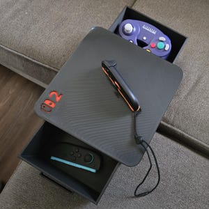 May include: A black gaming console table with a drawer, featuring a Switch logo in red. The table holds a purple GameCube controller, a black and orange Switch controller, and a black and teal Switch controller.
