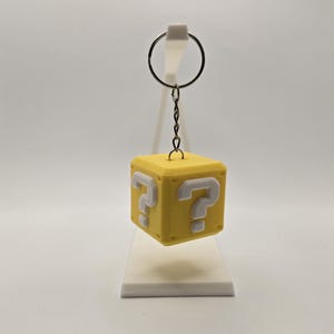 May include: A yellow cube-shaped keychain with white question marks on each side. The keychain is attached to a metal ring with a small chain. The item is a novelty item.