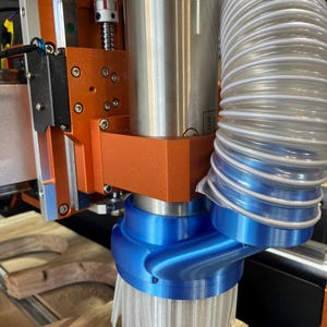 May include: Close-up of a CNC router with a blue dust collection shroud and a white brush attached to the spindle. The router is mounted on an orange base and has a silver metal spindle.