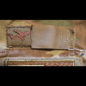 May include: A brown patch with a copper-coloured embroidered symbol of a stylised aircraft. The patch is attached to a brown fabric with a white stripe pattern.