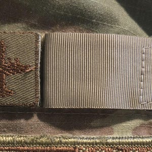 Puede incluir: Un parche marrón con un diseño de avión marrón cosido en una tela de camuflaje. Una correa de tela beige está cosida junto al parche.