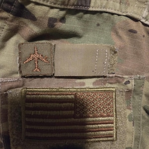 Op de afbeelding: Een camouflage militaire uniform met een bruine vliegtuigpatch, een bruin-witte Amerikaanse vlagpatch en een bruine cirkelpatch met de tekst "READY TO FLY".