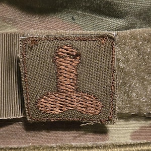 May include: A brown patch with a stylized brown figure on a camouflage fabric background.
