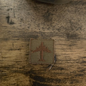 May include: A small, green patch with a brown embroidered image of an aeroplane. The patch is made of a woven fabric.