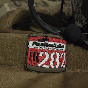 May include: A red and white embroidered patch with the text "IR282" and the words "Nuclear Sub" in a white font. The patch is attached to a brown fabric background.