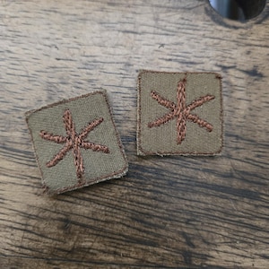 Könnte beinhalten: Zwei braune gestickte Aufnäher mit einem sechszackigen Sternmuster auf grünem Hintergrund.