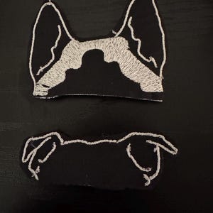 May include: Two black fabric patches with white embroidery depicting a dog's head. The top patch shows the dog's ears and the top of its head. The bottom patch shows the dog's nose and mouth.