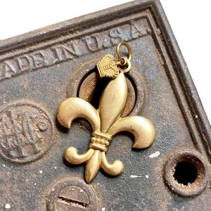 May include: Gold-toned fleur-de-lis charm with a small crown on top. The charm is sitting on a rusty metal surface with the words "MADE IN U.S.A." visible.
