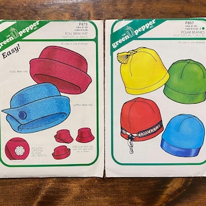 May include: Two vintage sewing pattern envelopes from "The Green Pepper, Inc." The patterns feature illustrations of hats, including roll brim hats and polar beanies, in various colors like red, blue, yellow, and green. The text on the envelopes includes "Easy!" and pattern details.