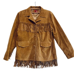 May include: A tan suede jacket featuring long fringe detailing across the chest and along the bottom hem. The jacket includes a collared neckline, two front pockets with flaps, and two central buttons. The interior label reads HAYASHI TAILOR, LTD.