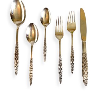 Puede incluir: Un juego de seis cubiertos plateados, que incluye cucharas, tenedores y un cuchillo. Los mangos presentan un patrón decorativo repetido. Los utensilios están dispuestos sobre un fondo blanco, mostrando sus superficies reflectantes.