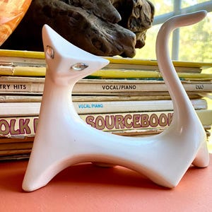 Puede incluir: Una figura de gato de cerámica blanca con un diseño elegante y moderno. El gato tiene una cola larga y curvada y detalles de cristal en los ojos. La figura se exhibe sobre una superficie coral, con partituras vintage de fondo.